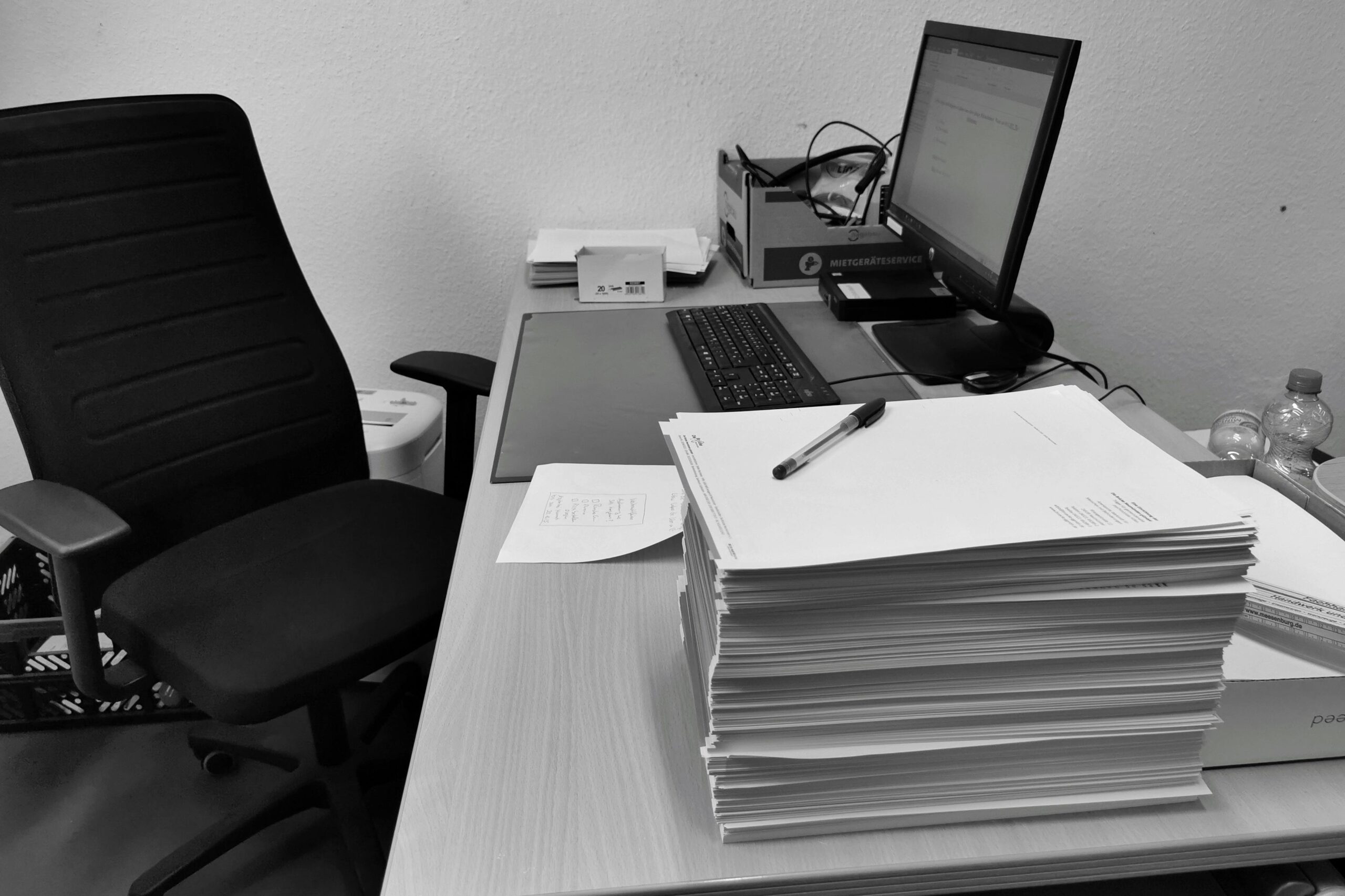 Black and white image of a cluttered office desk with a computer, chair, and paperwork.
