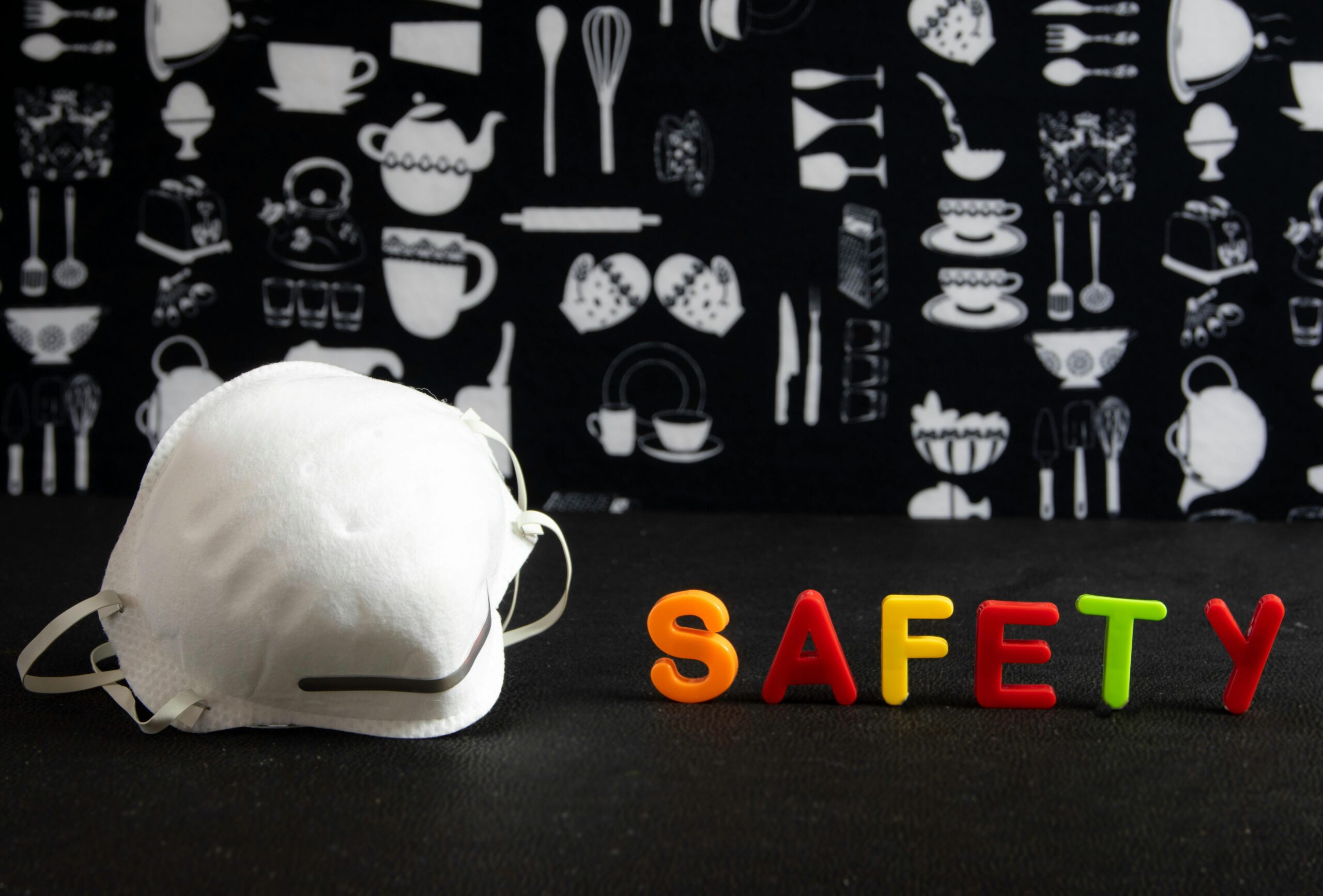 Face mask next to colorful 'SAFETY' letters against a kitchen-themed background.
