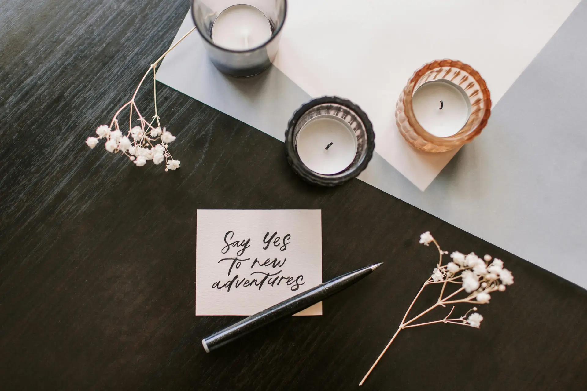 Flat lay of a motivational note, candles, flowers, and a pen on dark wooden table.
