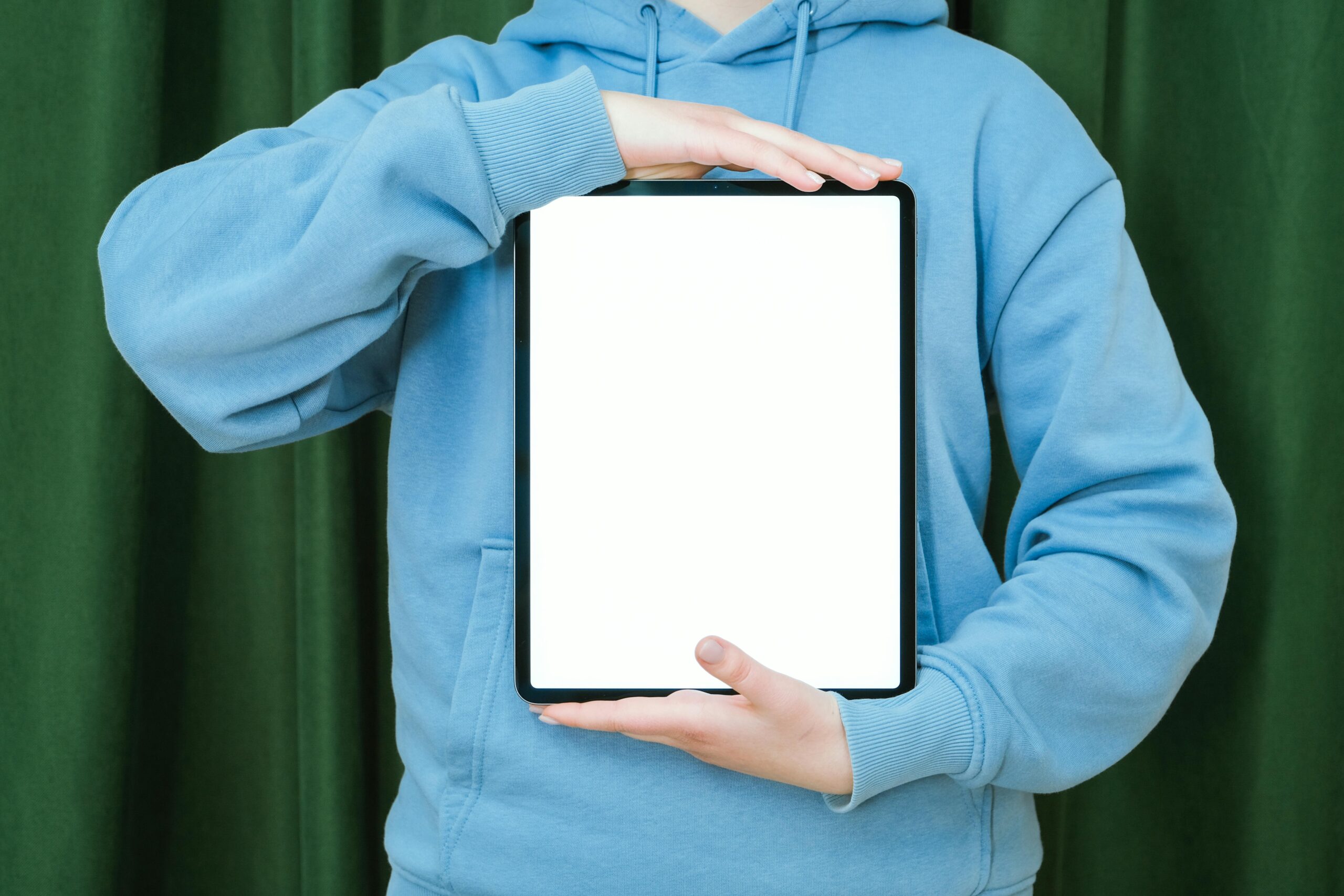 Person in a blue hoodie holding a blank tablet against a green curtain background.