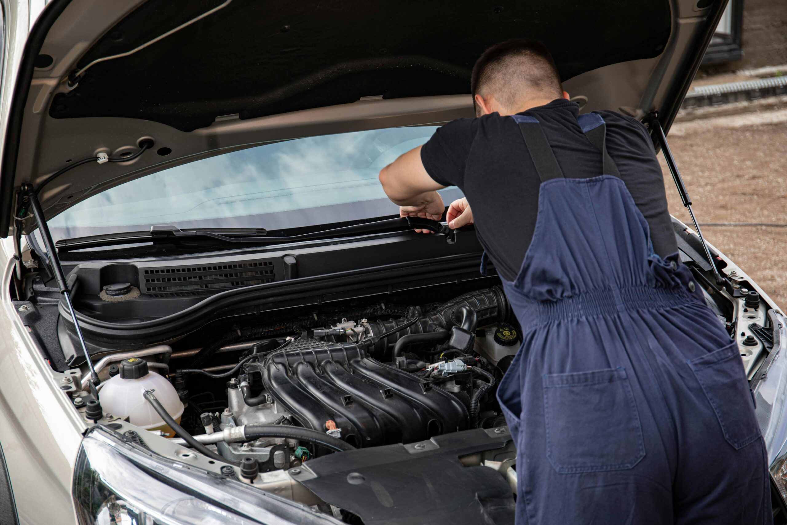A mechanic in blue overalls working on a car engine as part of maintenance routine.